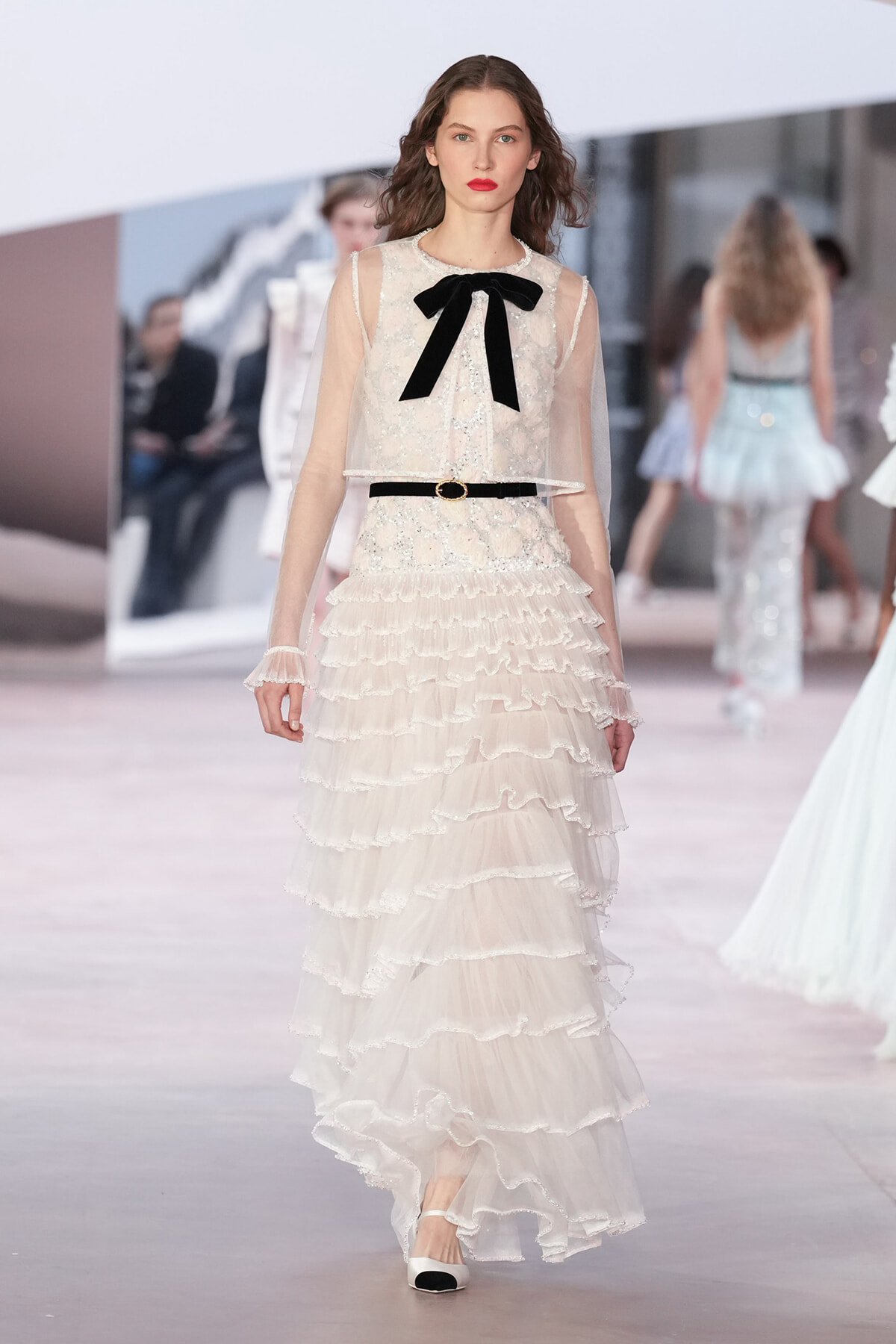 Runway model in a ivory lace gown with sheer sleeves, tiered ruffles, a slim belt, and a black bow at the neckline.