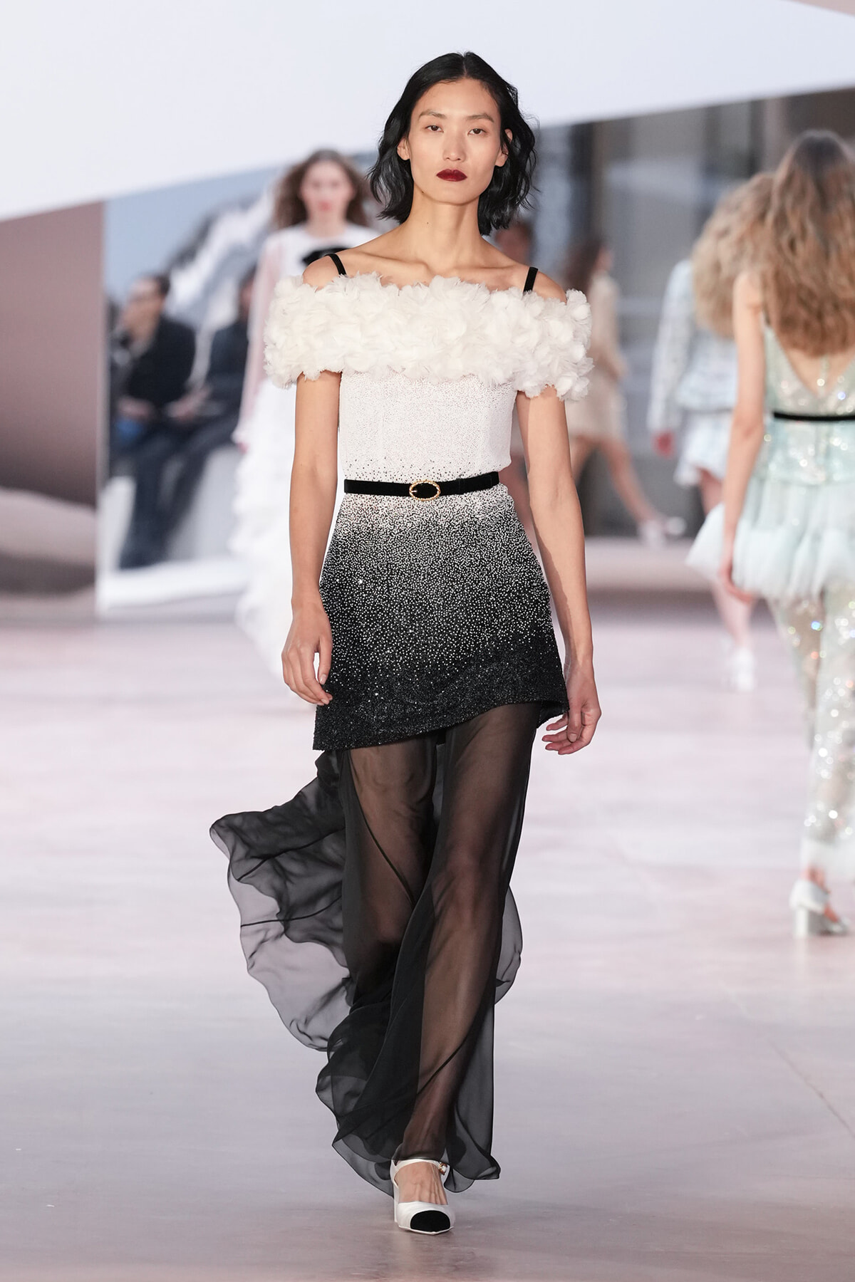 Model walking runway in off-shoulder white feathered top and gradient black-to-silver mini with sheer black overskirt, cinched with a black belt.