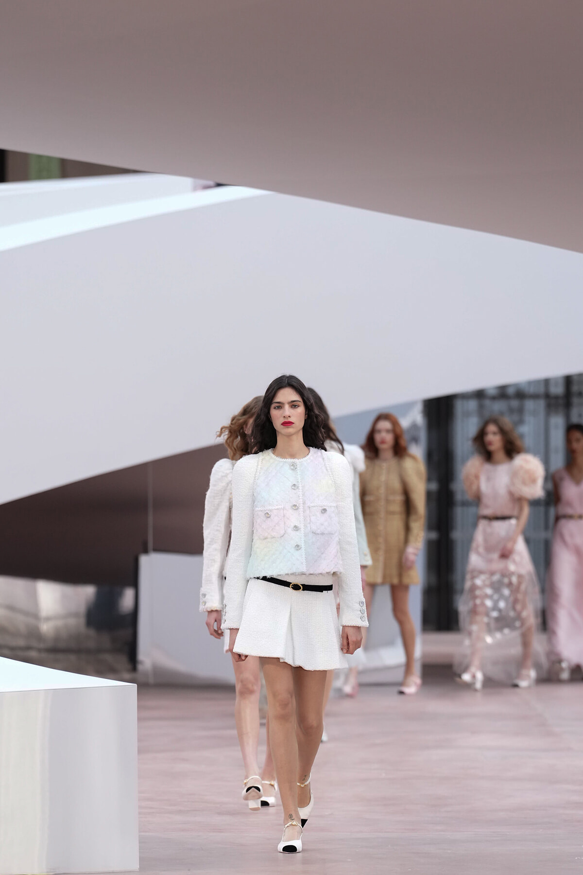 Model in white textured blazer and pleated skirt walks the runway at a fashion show, belt at waist, with other models in pastel outfits in the background.