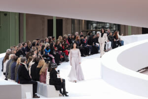 Model walks a white, curved runway as a spectator-filled audience watches a fashion show in an airy venue.