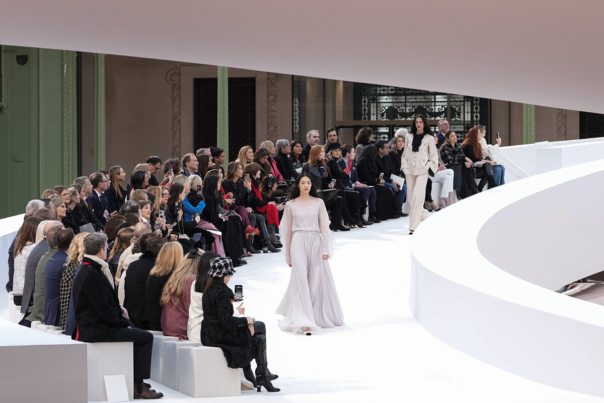Model walks a white, curved runway as a spectator-filled audience watches a fashion show in an airy venue.