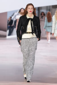 Female model walking a fashion show in a black textured blazer over a cream blouse with a black neck bow, and patterned wide-leg pants.