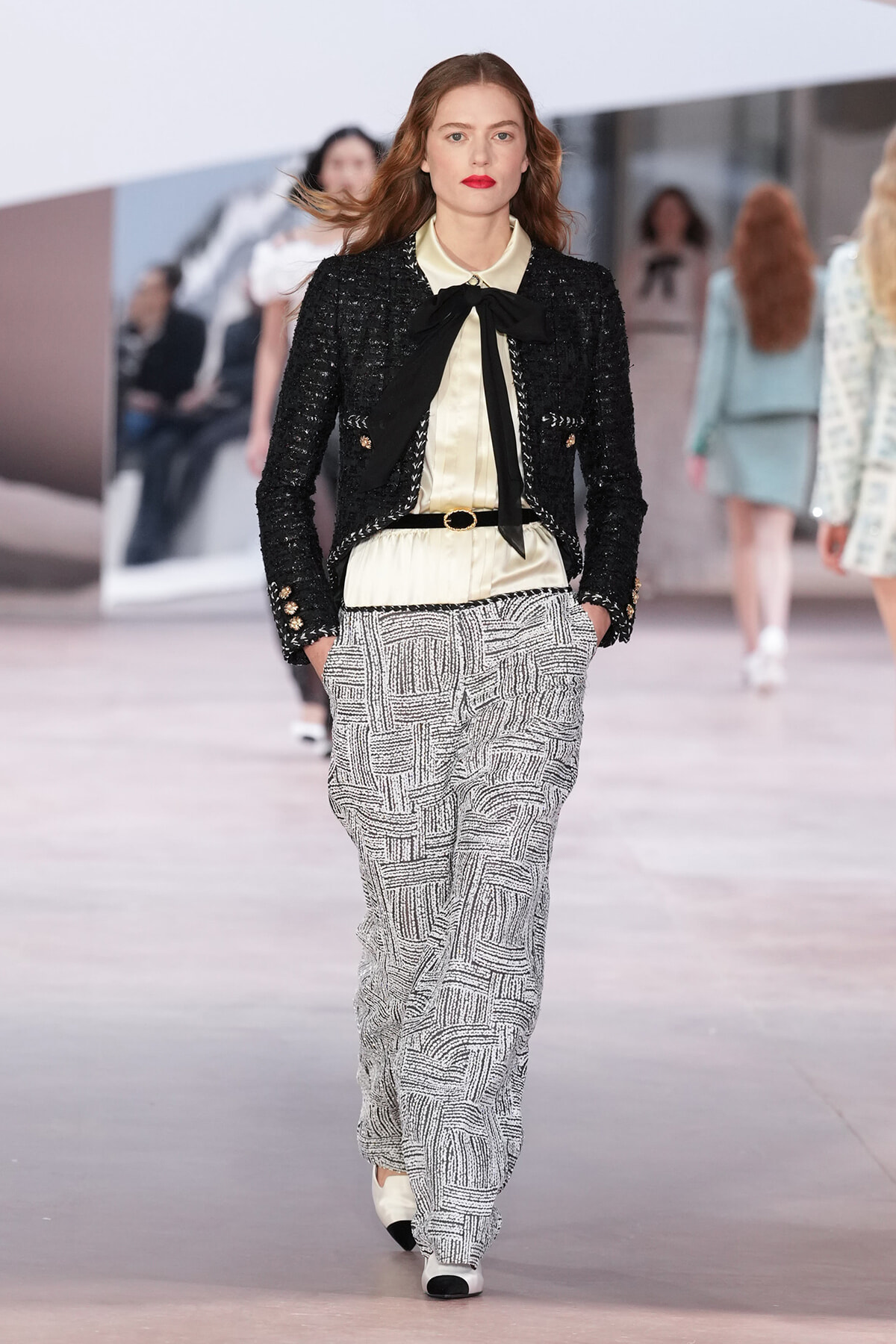 Female model walking a fashion show in a black textured blazer over a cream blouse with a black neck bow, and patterned wide-leg pants.