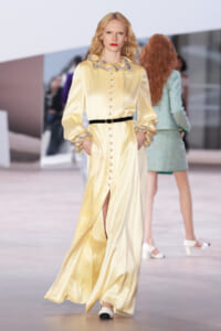 Model walks the runway in a pale yellow satin evening gown with a lace-edged collar, buttoned front, and a black belt.