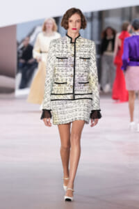 Model walks the runway in a cream tweed jacket and matching mini skirt with black piping and gold buttons, white heels.