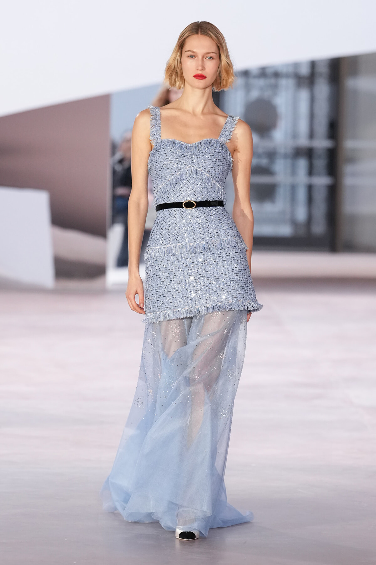 Model on a runway wearing a pale blue fringe-touched tweed dress, cinched with a black belt and a sheer glittery overskirt.