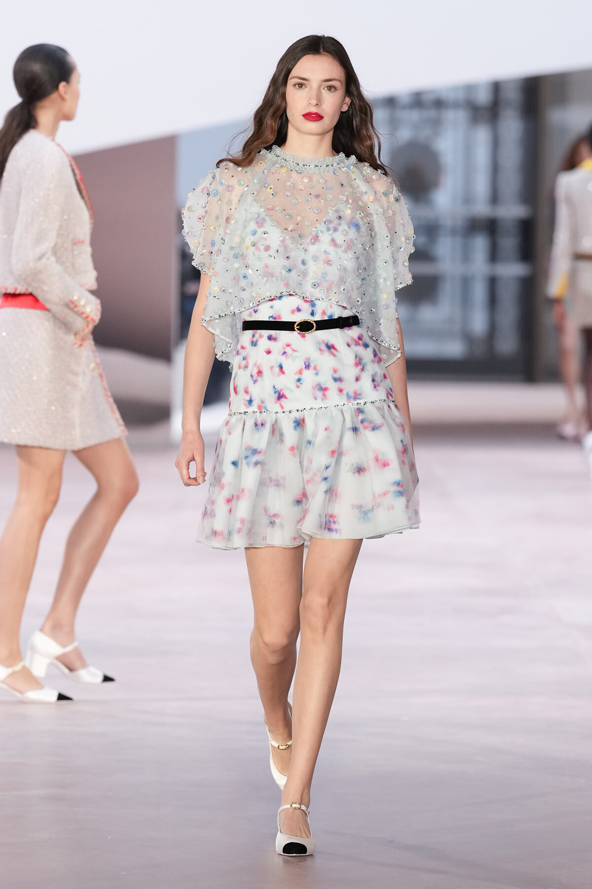 Model walks the runway in a pale floral mini dress with a sheer beaded top and a black belt, white heels.