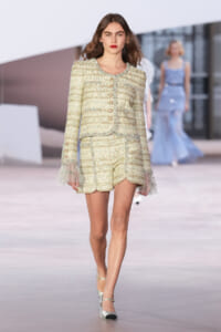 Model walking a runway in a pale tweed blazer and matching shorts with lace-trimmed cuffs and gold buttons.