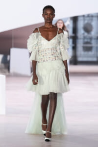 Runway model wearing a pale mint off‑the‑shoulder gown with ruffled sheer sleeves and a tiered tulle skirt.