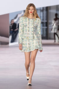 Model walking the runway in a white tweed ensemble with multicolored embellishments and matching jacket and mini skirt, white black‑toe heels.