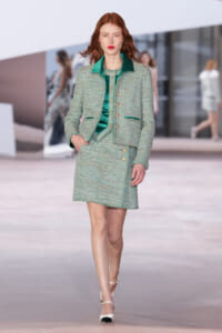 Model with red hair walks a runway wearing a mint-green tweed jacket and matching skirt, with a green satin blouse and gold buttons.