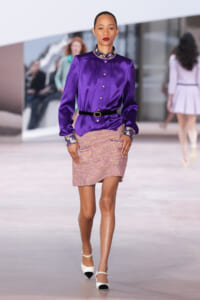 Model on runway wearing a purple satin blouse with a jeweled collar and cuffs, a black belt, and a pink tweed mini skirt with pockets and scalloped edge.