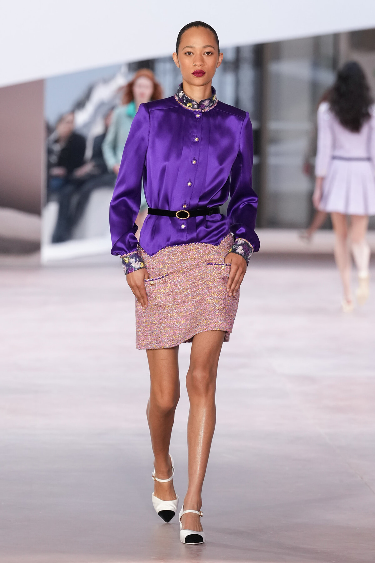 Model on runway wearing a purple satin blouse with a jeweled collar and cuffs, a black belt, and a pink tweed mini skirt with pockets and scalloped edge.