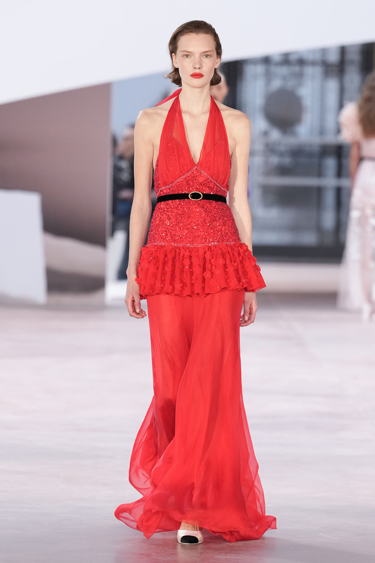 Model walks the runway in a red halter-neck sequin dress with a black belt and a layered ruffle peplum over a flowing chiffon skirt.