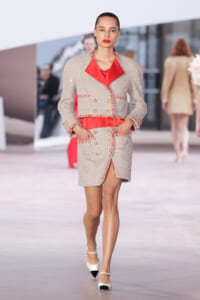 Model walking the runway in a beige tweed blazer and skirt with red satin blouse and red trim details