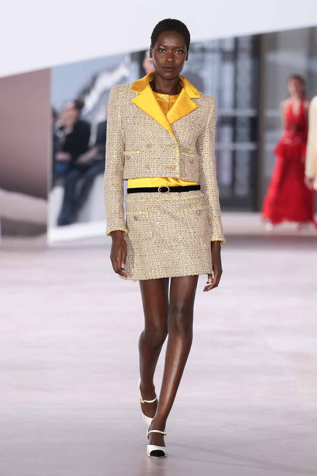 Model on a runway wearing a cream tweed blazer with bright yellow satin lapels and matching mini skirt, belted at the waist with a black belt.