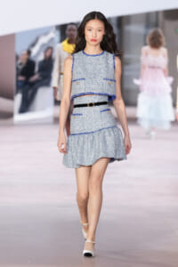 Female model walking a runway in a light blue tweed two-piece set with blue trim and a black belt.