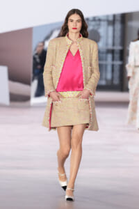 Model on the runway wearing a bright pink blouse with a cream tweed suit (jacket and mini skirt) and white heels, hands in pockets for a polished look.