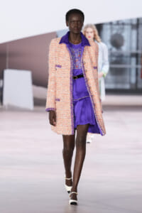 Female model walking the runway in a purple dress and peach textured coat during a fashion show, white heels visible on a spacious catwalk.