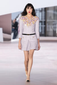 Model walking a runway in a pale lavender tweed dress with a multicolored woven V-neck, black belt, and cream heels.
