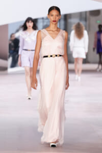 Model walks the runway in a pale pink satin sleeveless gown with a black belt and gold buckle, bold red lips.