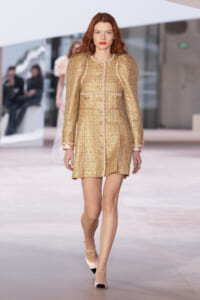 Model walks a runway in a gold textured mini dress with button front and puff sleeves, pink cuffs, and matching heels.