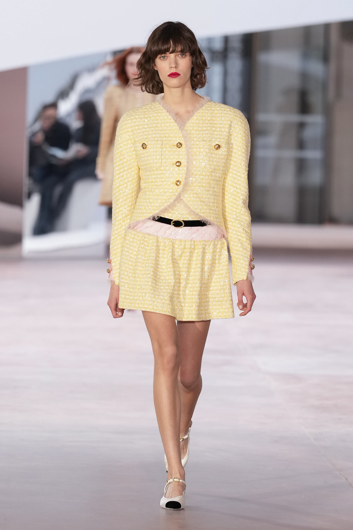 Model on a runway wearing a pale yellow tweed two-piece suit with gold buttons and a pink belt, paired with white shoes and red lipstick.
