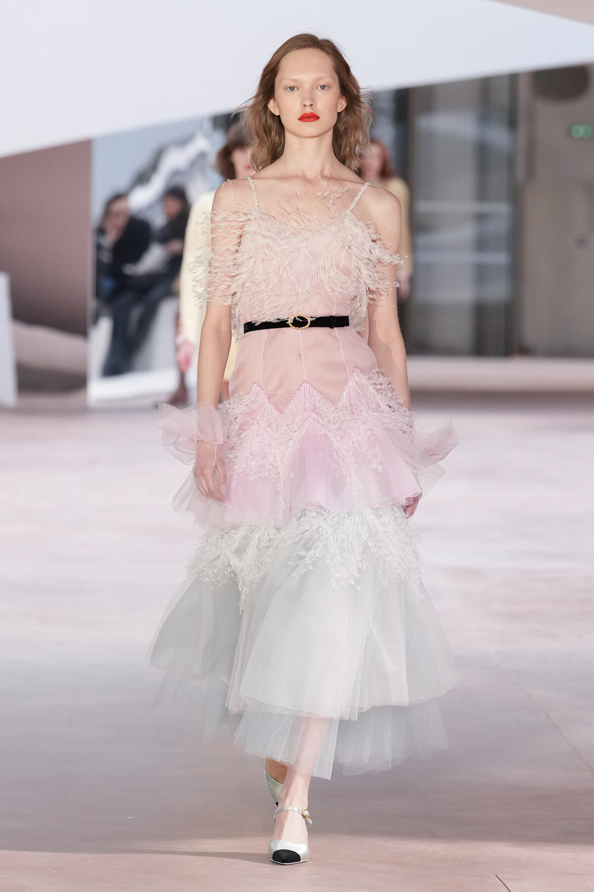 Runway model in a pale pink feathered gown with a black belt, walking a fashion show.