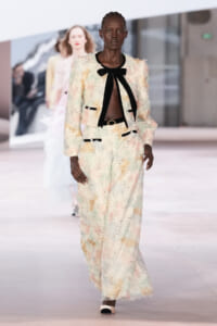 Model walks a pastel lace suit with a black bow tie, sheer top, and belt on a fashion runway