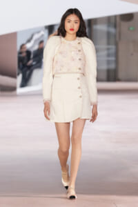 Model walks the runway in a cream textured two-piece suit: cropped jacket with gold buttons and a matching mini skirt with pearl trim and fringe details on cuffs and hem.