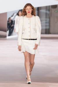 Model walking a fashion show in a white boucle jacket and matching mini skirt, accented with gold buttons and a thin black belt, wearing white heels.