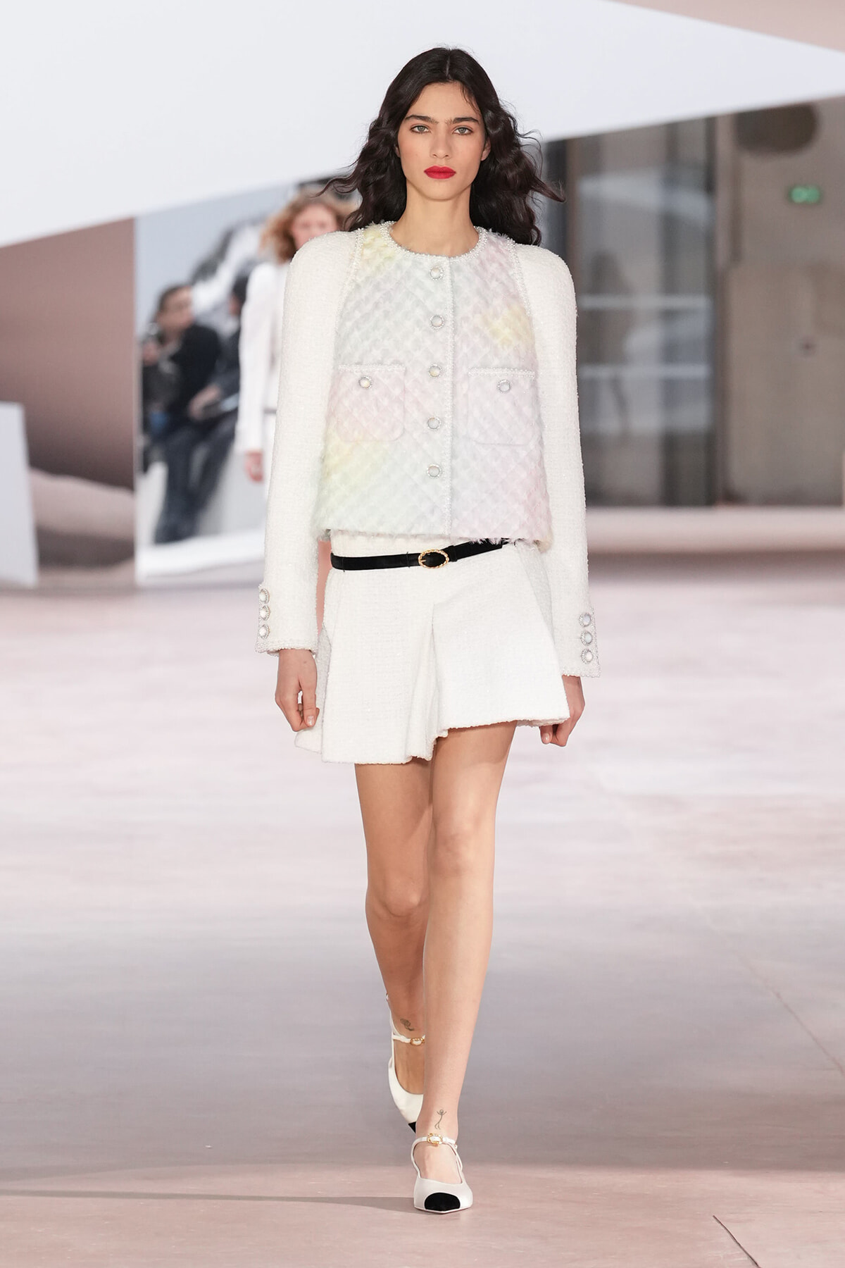Model walks a runway in a white textured jacket with pastel quilted front, matching white shorts, and a thin black belt.