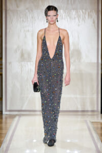 Model on a runway wearing a deep-plunge, jewel-studded gown covered in multicolored stones, holding a small black clutch.