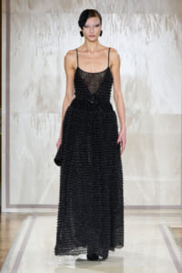 Model walking a runway in a floor-length black beaded gown with a sheer bodice and fringe details, wearing a black headpiece and matching accessories on a neutral backdrop.