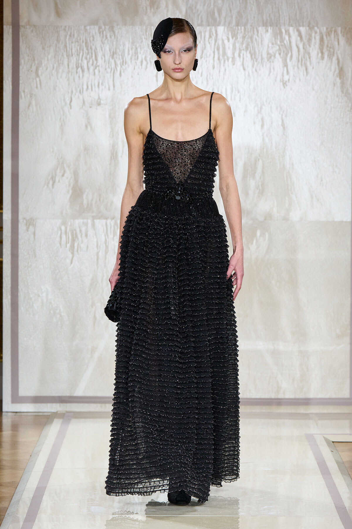Model walking a runway in a floor-length black beaded gown with a sheer bodice and fringe details, wearing a black headpiece and matching accessories on a neutral backdrop.