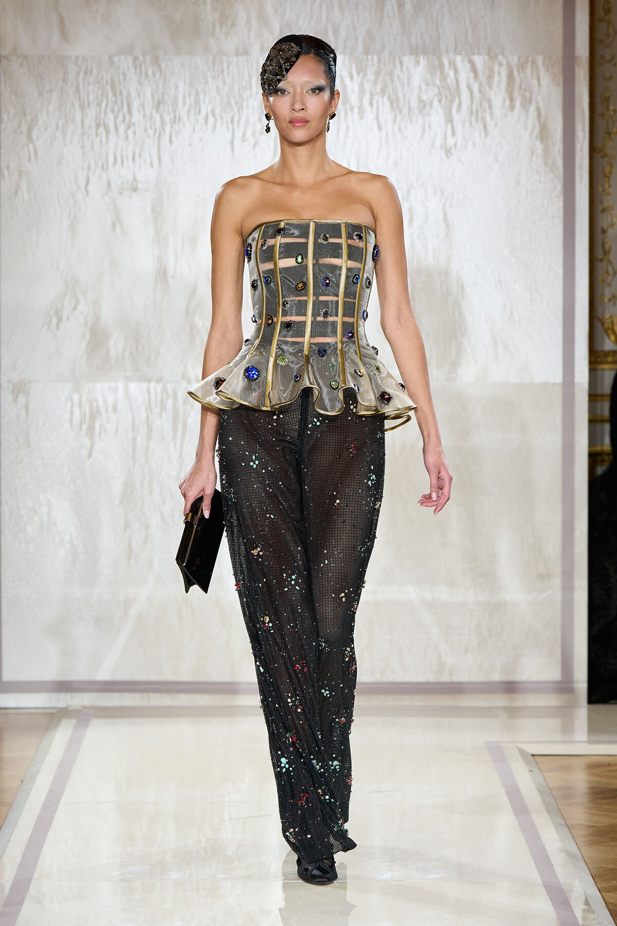 Model walks the runway in a strapless structured corset with a gold grid and jewel embellishments, paired with sparkly black sequined pants and a clutch.