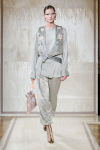 Model walking a fashion runway wearing a pale gray outfit with a sequined floral vest and matching pants, carrying a pink drawstring bag.