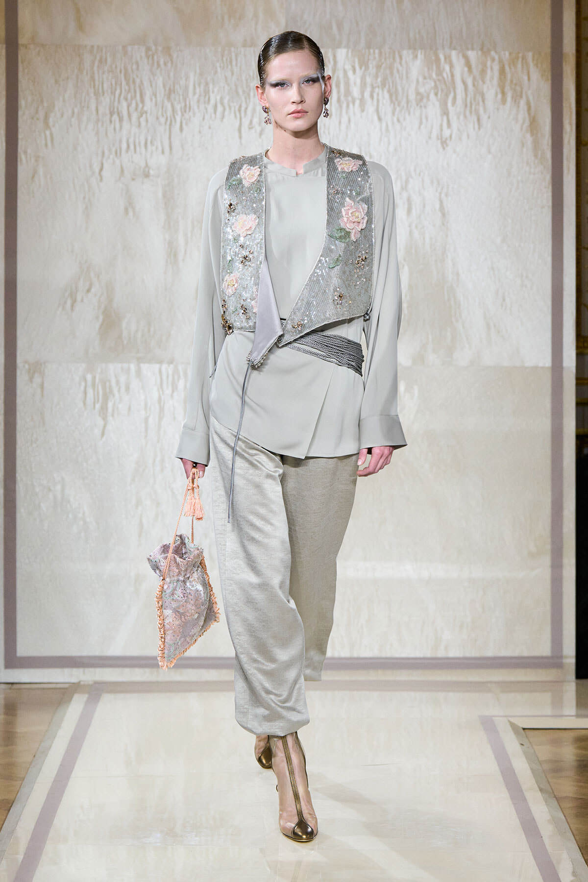 Model walking a fashion runway wearing a pale gray outfit with a sequined floral vest and matching pants, carrying a pink drawstring bag.