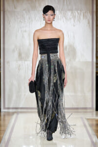 Runway model wears a black strapless top and a sparkling, beaded fringe skirt with hanging chains, holding a small black clutch.