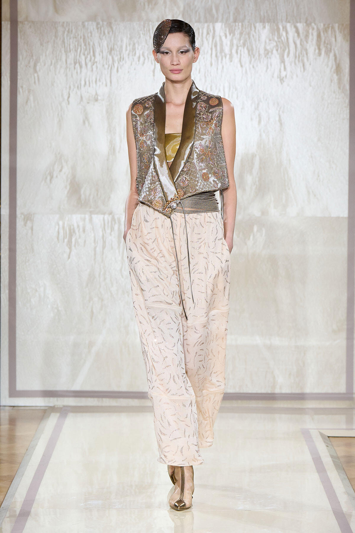 Model walks the runway in a metallic sleeveless vest with a draped front over a gold top, paired with pale patterned wide-leg pants and a textured belt.