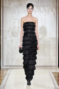 Model walking a runway in a strapless black tiered fringe gown, holding a small black clutch.