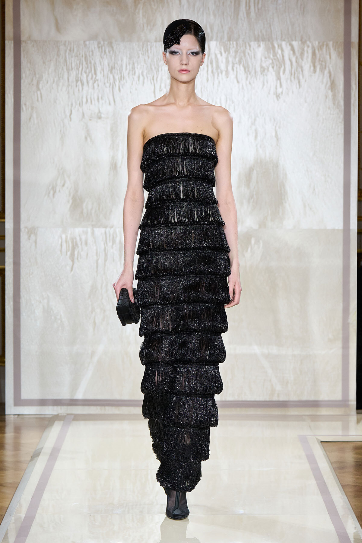 Model walking a runway in a strapless black tiered fringe gown, holding a small black clutch.