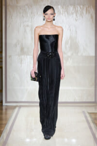 Model on a runway wearing a strapless black evening gown with a beaded waist brooch and a jeweled clutch.