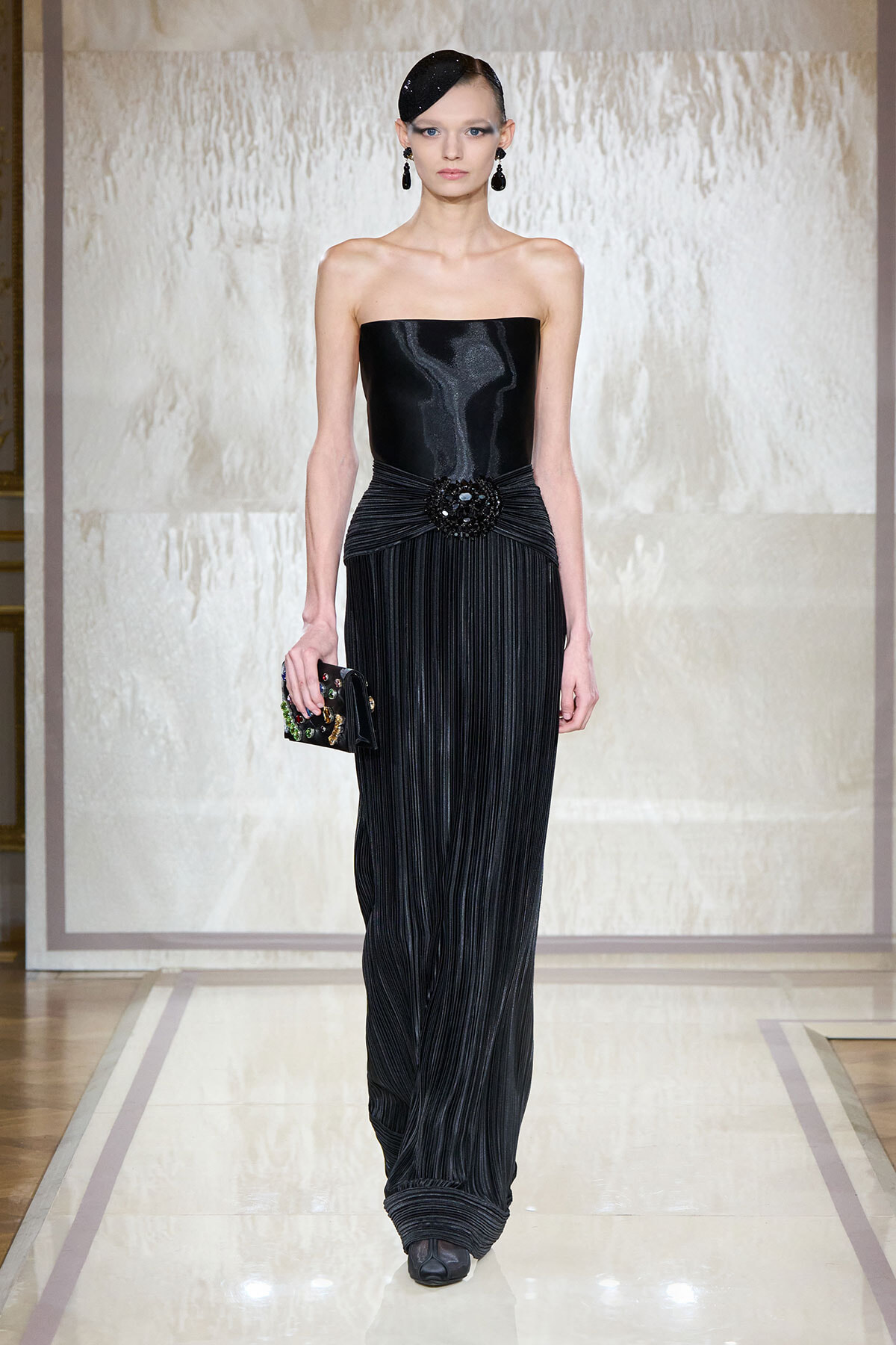 Model on a runway wearing a strapless black evening gown with a beaded waist brooch and a jeweled clutch.