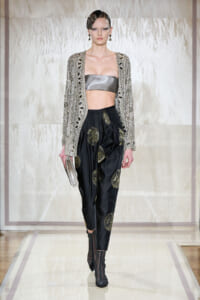 Runway look: metallic bandeau top, embellished cardigan, black satin pants with gold circular designs, ankle boots, silver clutch held in one hand.