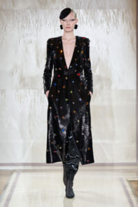 Model walks the runway in a long black sequined coat dotted with colorful gemstones and black boots.