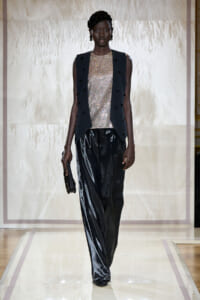 Model walking a runway in a metallic sequined top layered with a dark sleeveless vest and glossy black wide-leg pants, carrying a small black clutch.