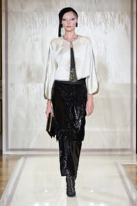 Model on a runway wearing a cream embroidered jacket over black sequined pants, black headscarf, and a black clutch.