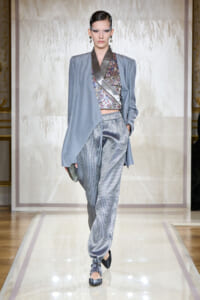 Model walking a runway in a pale blue draped jacket over a floral wrap top and metallic striped jogger-style pants, holding a small clutch.
