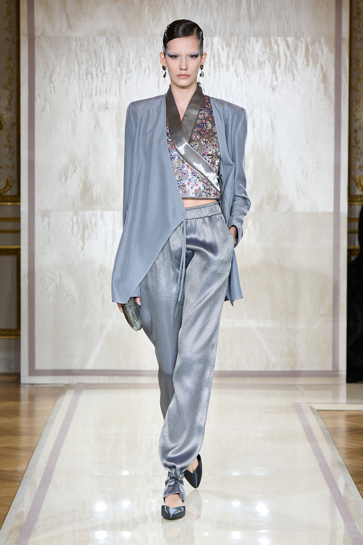 Model walking a runway in a pale blue draped jacket over a floral wrap top and metallic striped jogger-style pants, holding a small clutch.